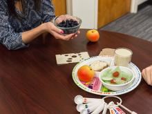 A plate of healthy foods like salad and apple, someone holding a bowl of blueberries, measuring spoons, bread, cheese and apple on a table.