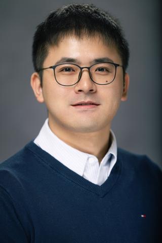 Man with glasses smiling, wearing a navy blue sweater and white shirt.