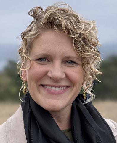 Jennifer Makelarski, smiling, with curly blonde hair pulled back, wearing a black scarf, with trees and sky in the background